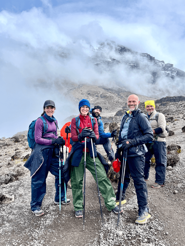 trekking kilimanjaro cume → Strike Tours Na imagem é possível observar um grupo de pessoas a sorrir para a fotografia, com bastões de caminhada, com o cume do Kilimanjaro atrás delas.