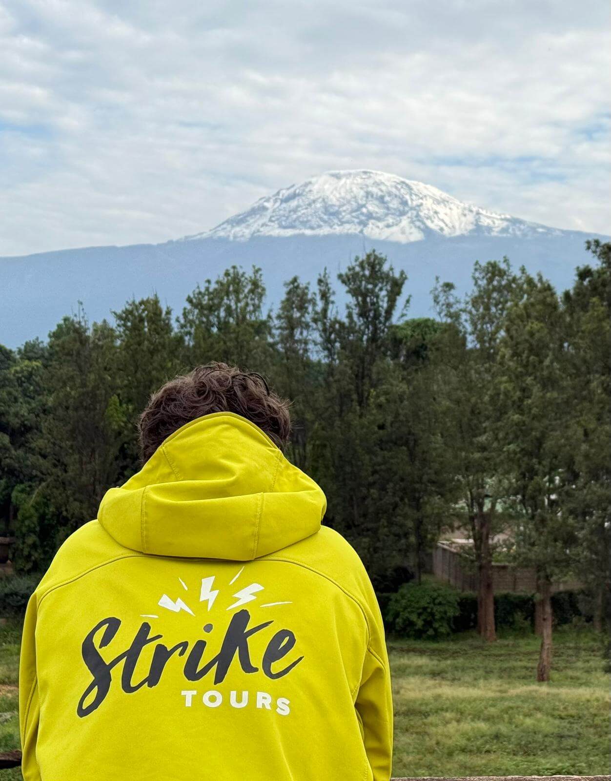 Person in a bright yellow hoodie with 'Strike Tours' on the back, standing outdoors with a snow-capped mountain in the distance.